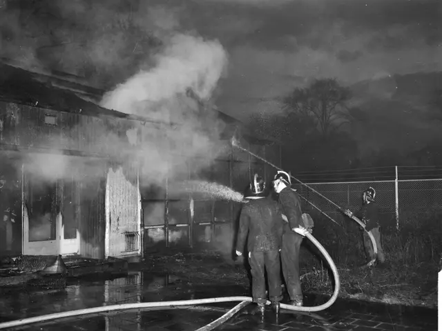 The image shows a group of firefighters wearing helmets and holding pipes, standing in front of a...