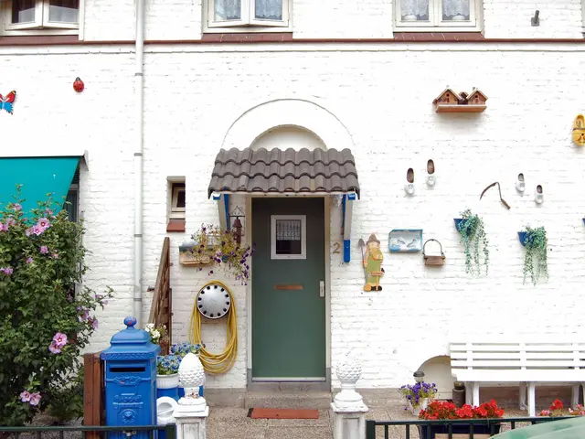The image shows a white house with a green door and windows, a bench in front of it, plants with...