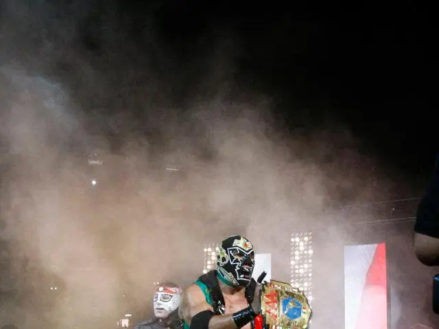 The image shows a man in a wrestling ring wearing a mask and holding a trophy, surrounded by a...