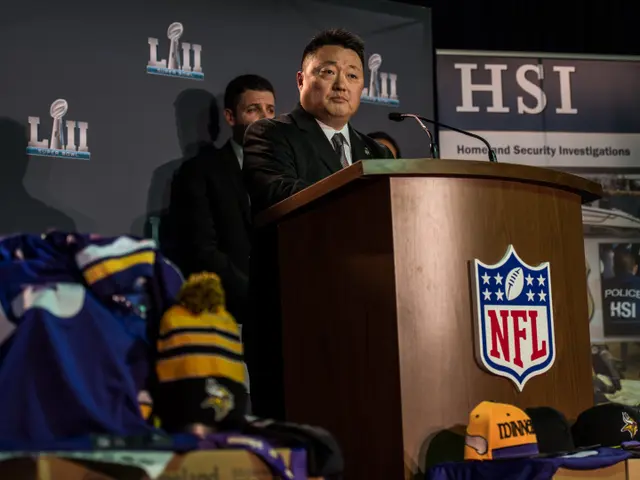 The image shows a man standing at a podium in front of a crowd. He is wearing a black blazer, white...
