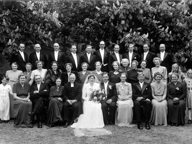 The image shows a black and white photo of a wedding party, with the bride and groom in the center,...