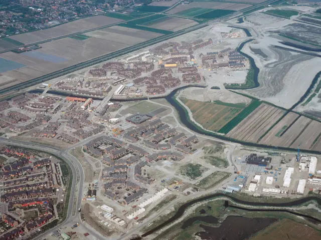 The image shows an aerial view of a city with lots of buildings, roads, fields, trees, and water....