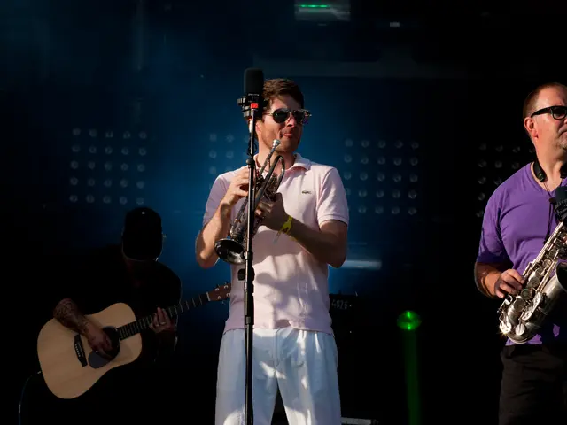 The image shows two men wearing goggles and playing saxophones on stage at a concert. There is a...