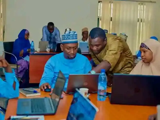 The image shows a group of people sitting around a table with laptops, bottles, cups, books, pens,...
