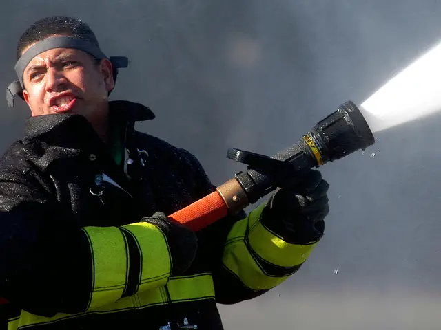 The image shows a firefighter wearing a black jacket, gloves, and a cap, holding a hose and...
