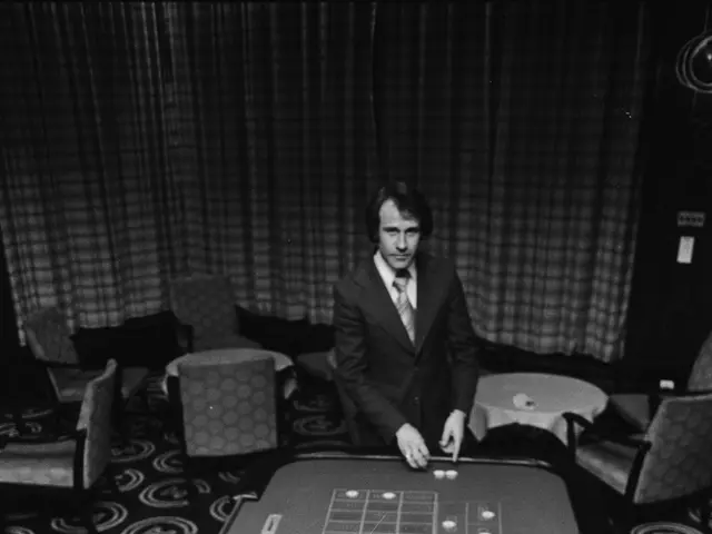 The image shows a man in a suit sitting at a roulette table in a casino. He is surrounded by chairs...