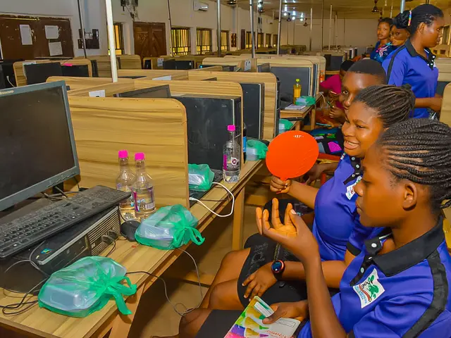 The image shows a group of young girls sitting at desks in front of computers, with monitors,...