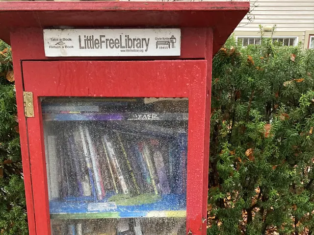 The image shows a red little free library in front of a house, surrounded by trees and leaves on...