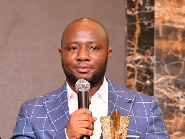 The image shows a man in a blue suit holding a microphone and a trophy, with a wall in the...