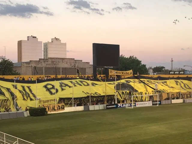 The image shows a soccer field with a group of people standing on top of it, surrounded by a metal...