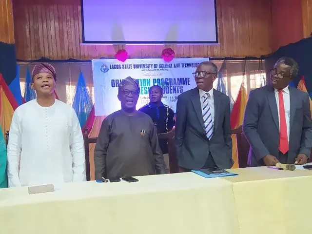 The image shows a group of people standing around a table with flower vases, books, and other...