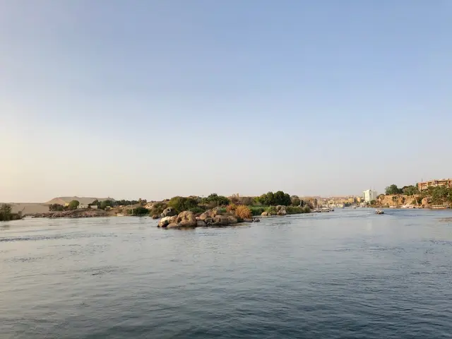 The image shows the Nile River in Egypt, with boats on the water, rocks, trees, buildings, and a...