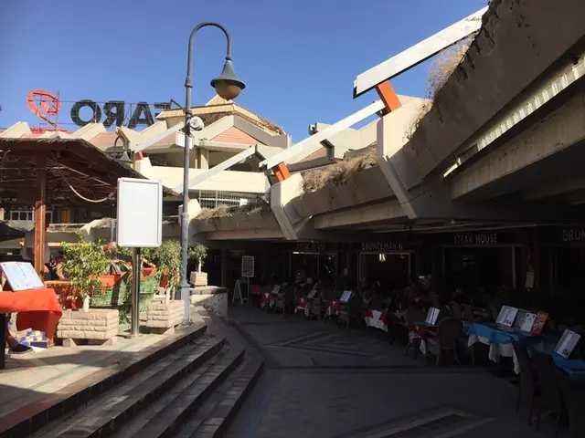 The image shows a street with tables and chairs in the middle of it, surrounded by buildings,...