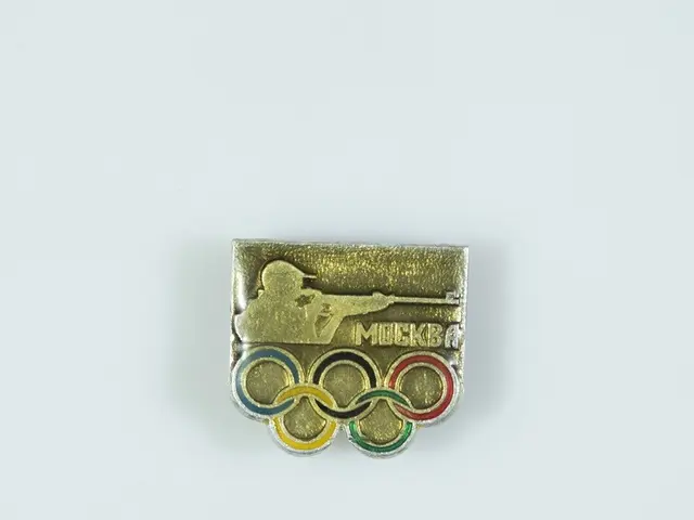 The image shows a gold and silver pin with the Olympic rings on it, set against a white background....
