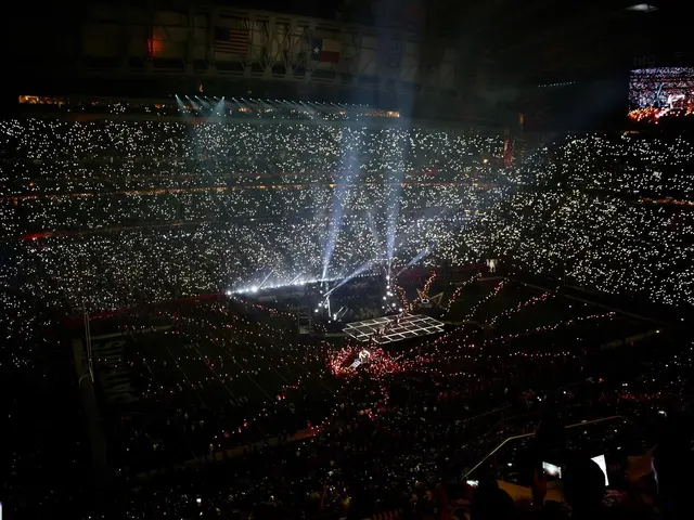 The image shows a large crowd of people in a stadium at night, illuminated by bright lights and...