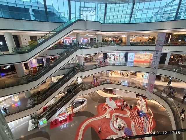 The image shows a large atrium filled with lots of people, escalators, railings, stores, lights,...