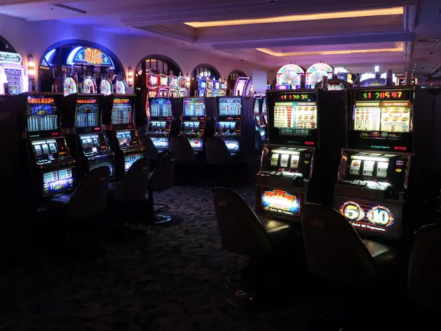 The image shows a row of slot machines in a casino, with chairs on the floor and lights on the...
